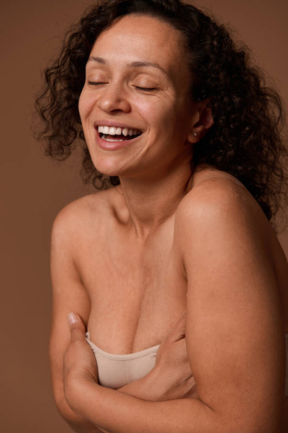Delighted confident dark-haired curly woman with closed eyes and skin flaws in beige underwear smiles toothy smile posing against beige background with copy space for ads - Photo, Image