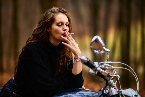 A beautiful long-haired woman smoking on a chopper motorcycle in autumn landscape on a forest road - Photo, Image