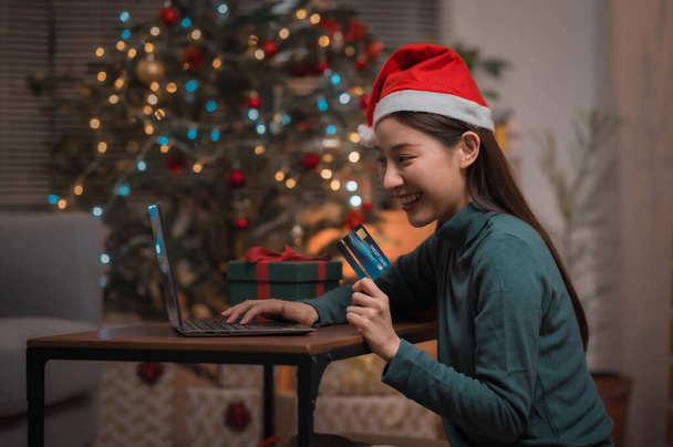 Boldog ázsiai nő mikulás kalap segítségével hitelkártya és laptop vásárlás során Christman nyaralás otthon - Fotó, kép