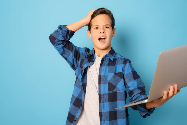 Kid boy shocked holding laptop computer standing over blue background. - Foto, Bild