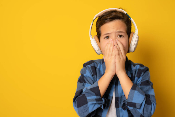 Маленький белый мальчик в наушниках, слушающий музыку на жёлтом фоне, шокирован, закрывая рот руками за ошибку. Секретная концепция. - Фото, изображение