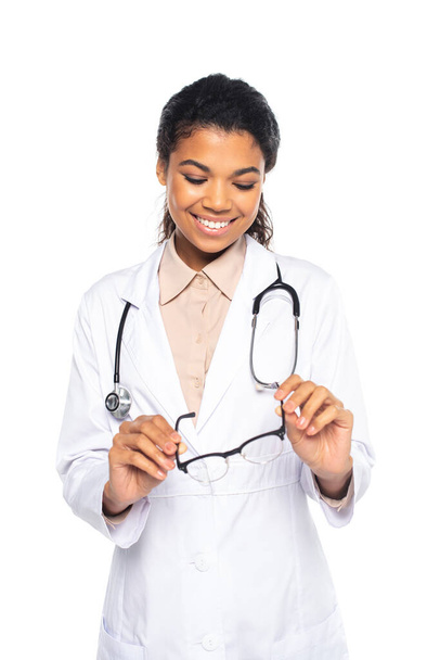 Smiling african american doctor holding eyeglasses isolated on white  - Photo, Image