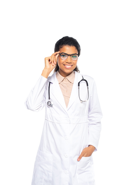 Young african american ophthalmologist holding eyeglasses and hand in pocket of white coat isolated on white  - Фото, изображение