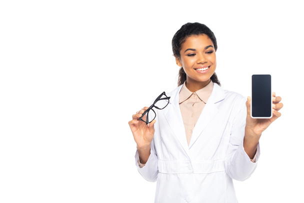 Positive african american ophthalmologist holding cellphone with blank screen and eyeglasses isolated on white  - Fotografie, Obrázek