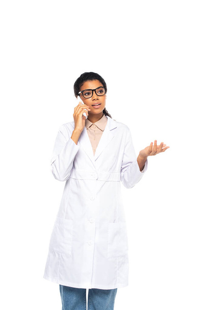 Молодой африканский американский врач в очках разговаривает на смартфоне, изолированном на белом  - Фото, изображение