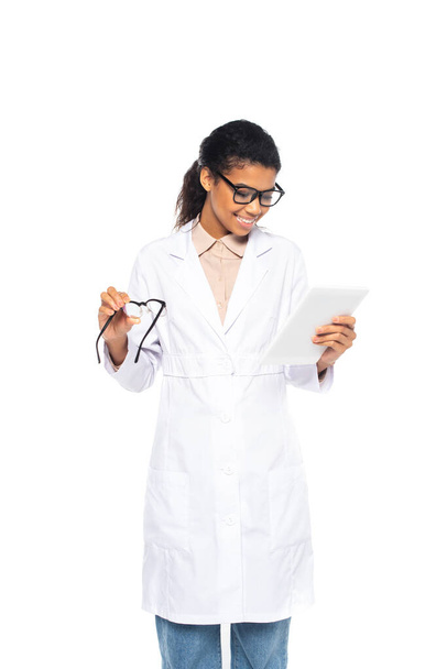 African american oculist in white coat holding digital tablet and eyeglasses isolated on white  - Foto, afbeelding