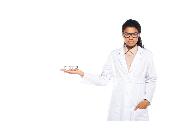 African american ophthalmologist holding eyeglasses and hand in pocket of white coat isolated on white  - Foto, imagen