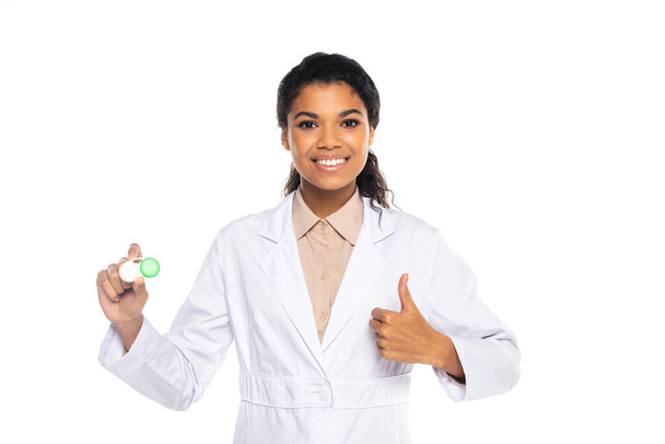 Smiling african american doctor in white coat holding container with lenses and showing thumb up isolated on white  - Foto, Bild