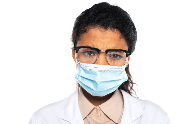Triste médico afroamericano en anteojos y máscara médica mirando a la cámara aislada en blanco  - Foto, imagen