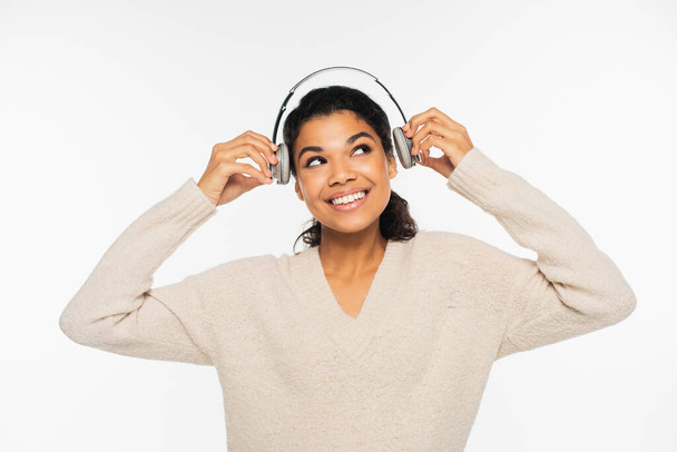 Cheerful african american woman in sweater holding headphones isolated on white - 写真・画像
