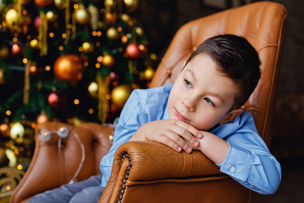 Счастливый ребенок сидит в кресле с украшением и елкой на заднем плане. Мальчик в голубой рубашке на фоне золотой рождественской елки отворачивается. - Фото, изображение