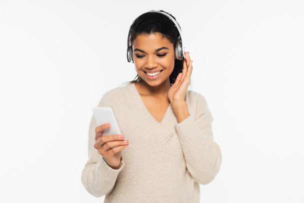Alegre mujer afroamericana en suéter usando auriculares y teléfonos celulares aislados en blanco - Foto, Imagen