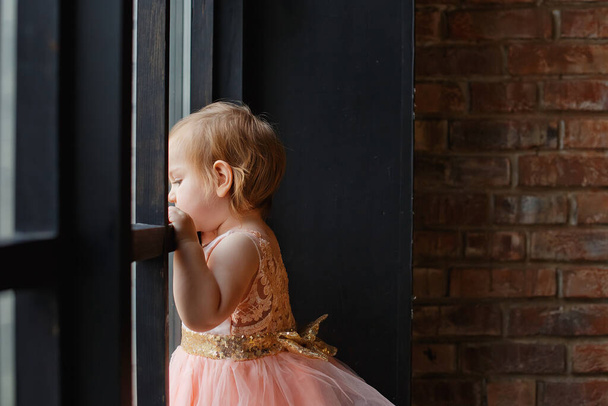 Une petite fille se tient près de la fenêtre sur le fond d'un mur de briques - Photo, image