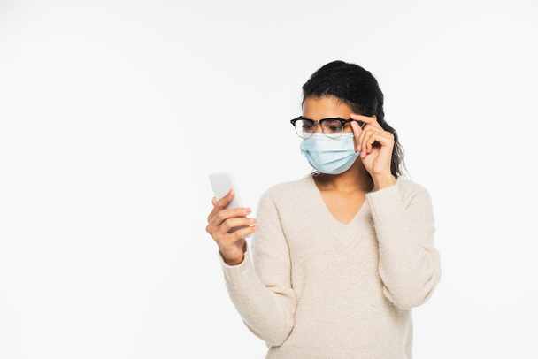 Junge Afroamerikanerin in medizinischer Maske und Brille mit Smartphone isoliert auf weiß - Foto, Bild