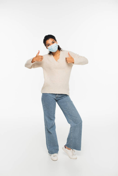 Full length of african american woman in medical mask showing thumbs up on white background  - Foto, Bild