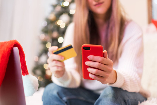 携帯電話、クレジットカード、ラップトップを持つ女性の閉鎖。クリスマス休暇でオンラインショッピング。家からの女の子のテキストメッセージ。メリークリスマスとクリスマスツリーと幸せな新年 - 写真・画像