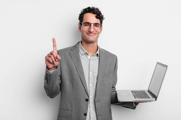 hispanic handsome man smiling and looking friendly, showing number one. business and laptop concept - Photo, image