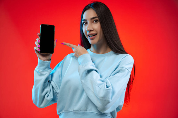Cropped portrait of young joyful girl pointing at phone screen isolated over red background - Фото, изображение