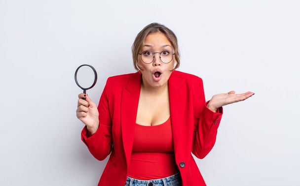 hispanic pretty woman looking surprised and shocked, with jaw dropped holding an object. magnifying glass concept - Фото, зображення