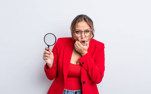 hispanic pretty woman with mouth and eyes wide open and hand on chin. magnifying glass concept - Photo, image