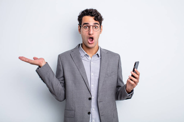 hispanic handsome man looking surprised and shocked, with jaw dropped holding an object. business and phone concept - Φωτογραφία, εικόνα