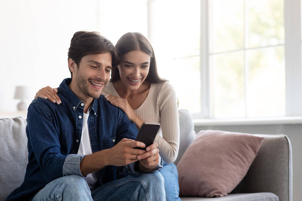 Smiling young caucasian man and woman looking at smartphone together, have video call - Fotografie, Obrázek