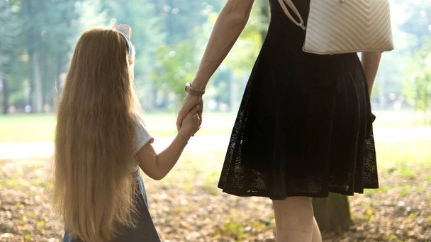 Back view of mom and her little daughter walking together holding hands in summer park - Zdjęcie, obraz