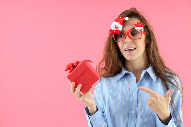 Aantrekkelijke vrouw in kerstbril houdt geschenkdoos op roze achtergrond - Foto, afbeelding