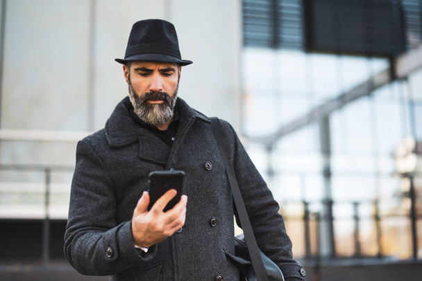 Ritratto di uomo d'affari in abiti neri in piedi di fronte all'edificio aziendale e guardando lo smartphone. Uomo barbuto nero sta utilizzando smartphone. - Foto, immagini