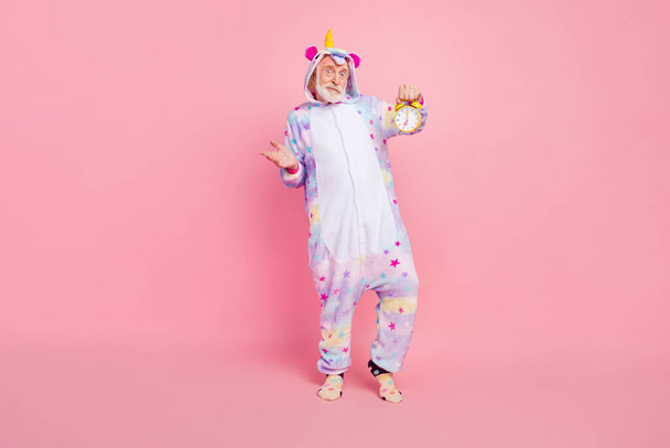 Photo of cute doubtful man pensioner unicorn sleepwear holding watch shrugging shoulders isolated pink color background - Fotó, kép