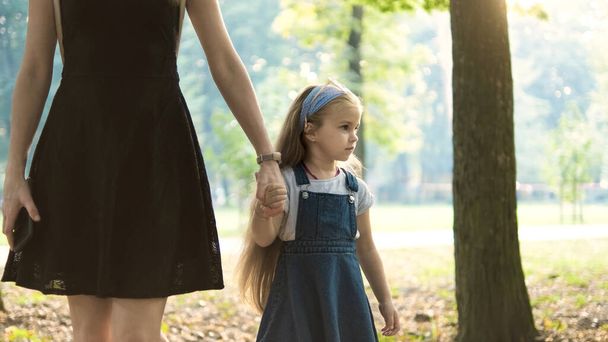 Anya és a kislánya hosszú hajjal, kéz a kézben sétálnak a nyári parkban. - Fotó, kép