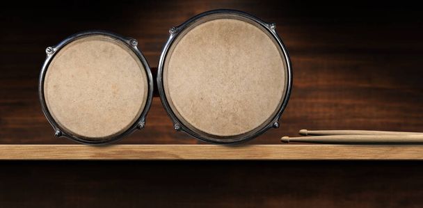 Großaufnahme von Bongo-Trommeln und einem Paar Drumsticks, über einem Holzregal mit Kopierraum. Schlaginstrument. Fotografie. - Foto, Bild
