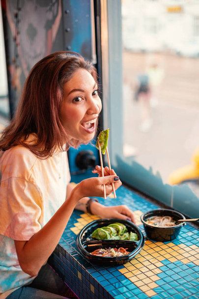 Gülümseyen Asyalı kadın pencerenin yanındaki fast food restoranında japon yeşili hamur tatlısı yiyor Jiaozi veya Gyoza - Fotoğraf, Görsel