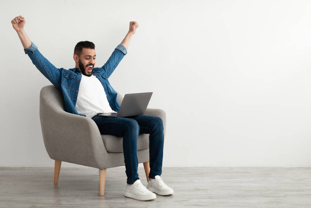 Jeune homme arabe excité assis dans un fauteuil avec ordinateur portable, célébrant le succès du travail, gagner à la loterie en ligne, espace de copie - Photo, image
