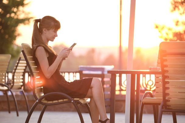 Eine junge Frau sitzt an einem Straßencafé-Tisch und schmökert an einem warmen Sommerabend im Freien mit ihrem Handy. Kommunikations- und mobiles Verbindungskonzept - Foto, Bild