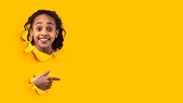 Surprised cheerful african american teenager girl pupil showing finger at empty space looks through hole on yellow paper - Foto, Imagem