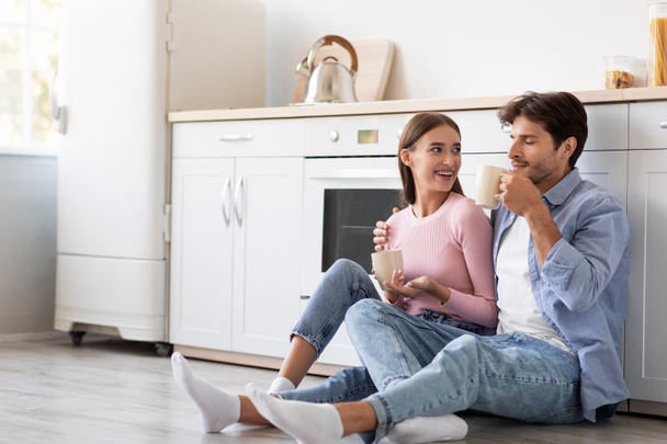 Satisfied glad young european lady and guy with cups of favorite drink sits on floor, talk in kitchen - Photo, Image