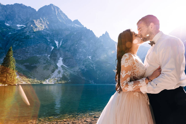 una chica joven en un vestido de novia con un chico en un traje, tomados de la mano, besándose en un camino de piedra a lo largo del lago. - Foto, Imagen
