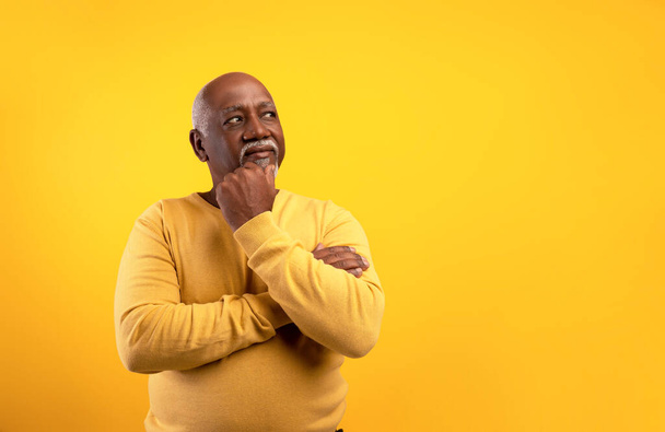 Задумчивый старший черный мужчина смотрит в сторону на пустое пространство, трогает подбородок, глубоко в мыслях на оранжевом фоне - Фото, изображение