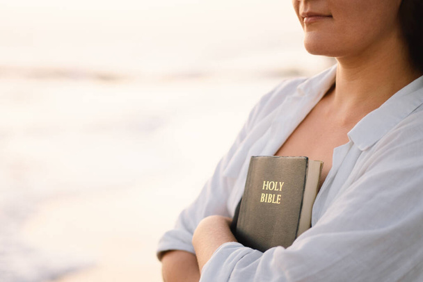 Христианка держит Библию в руках. Чтение Библии на море во время прекрасного заката - Фото, изображение