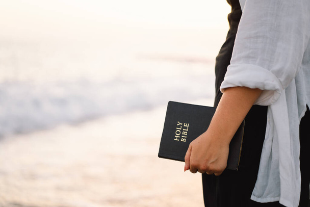 Христианка держит Библию в руках. Чтение Библии на море во время прекрасного заката - Фото, изображение