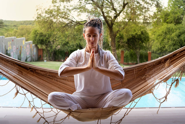 Blond yogi vrouw met dreadlocks in gebed positie zittend in een geweven hangmat. - Foto, afbeelding