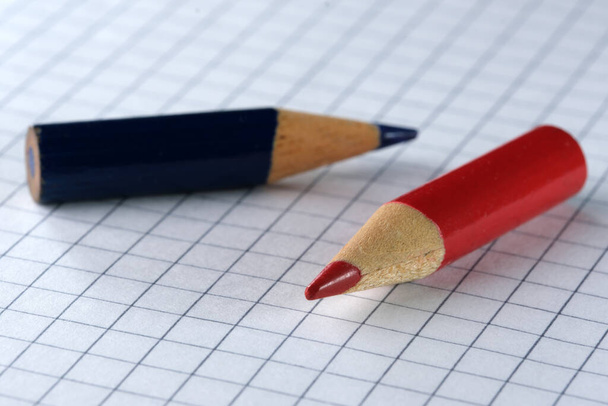 Stare małe używane naostrzone ołówki z czerwonego i niebieskiego drewna łupkowego leżą na kartce papieru w kratkę, zbliżenie - Zdjęcie, obraz