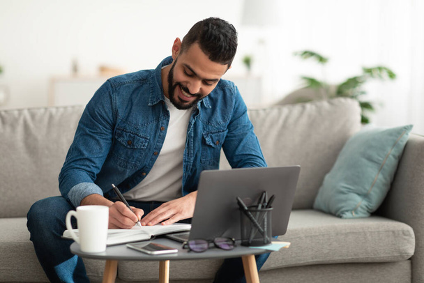Щасливий молодий арабський чоловік робить нотатки під час роботи на ноутбуці вдома, копіюйте простір. - Фото, зображення