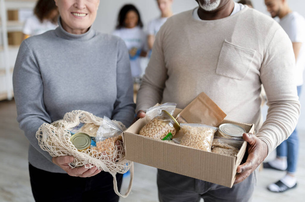 Cónyuges de edad avanzada de raza mixta mostrando comida de donación en caja y bolsa de cuerda, voluntarios empacando cajas para personas pobres - Foto, imagen