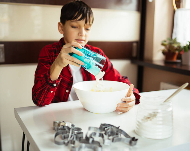 teenage boy sits at table in home kitchen and pours flour from glass into bowl to prepare dough for baking. Cookies are made at home, hobby, teenager 11 years old, lifestyle. - Foto, Imagem