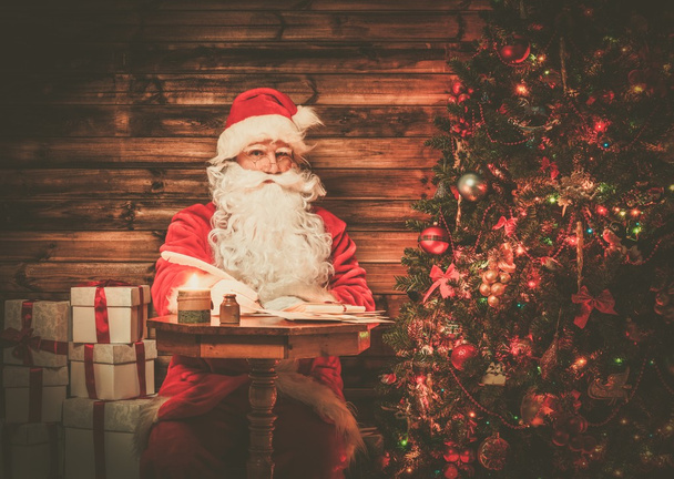 Santa Claus en el interior de la casa de madera sentado detrás de la mesa y escribir cartas con pluma de pluma
 - Foto, Imagen