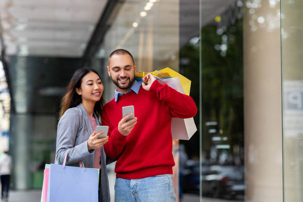 Young interracial couple with smartphones standing on street with shopper bags, buying goods online near big city mall - Photo, image