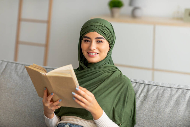Άνετο μείνετε στο σπίτι χόμπι. Νεαρή Άραβα γυναίκα σε hijab ανάγνωση βιβλίο, κάθεται στον καναπέ στο σαλόνι - Φωτογραφία, εικόνα