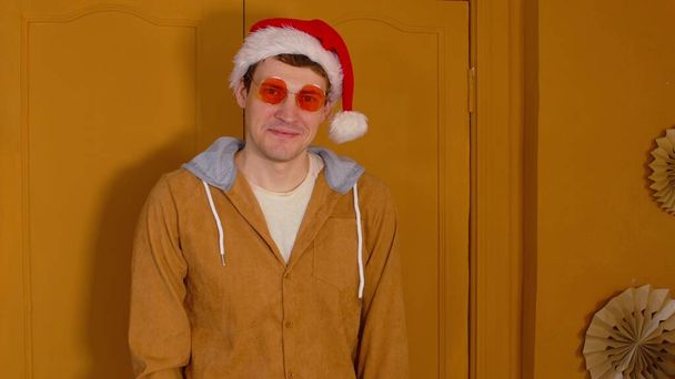 Happy man in santa hat and fashionable glasses expresses positive and festive emotions on brown background. Stylish male looking at camera and smiling, standing in room. - Foto, immagini